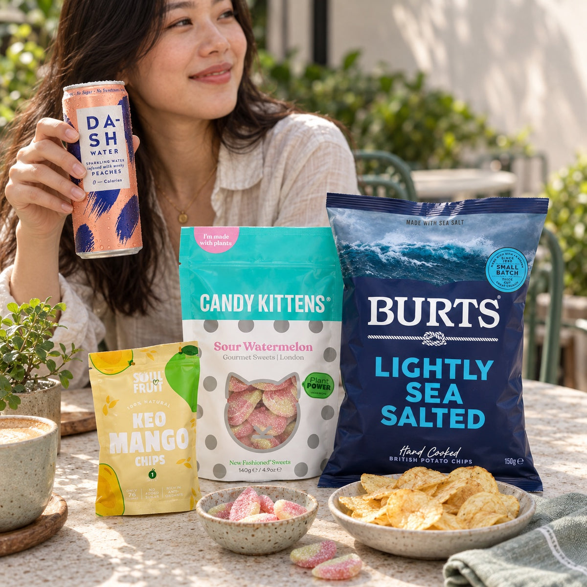 A woman enjoying DASH Peach sparkling water at a sunny café table with Burts Lightly Sea Salted crisps, Candy Kittens Sour Watermelon sweets and Soul Fruit Keo Mango Chips.